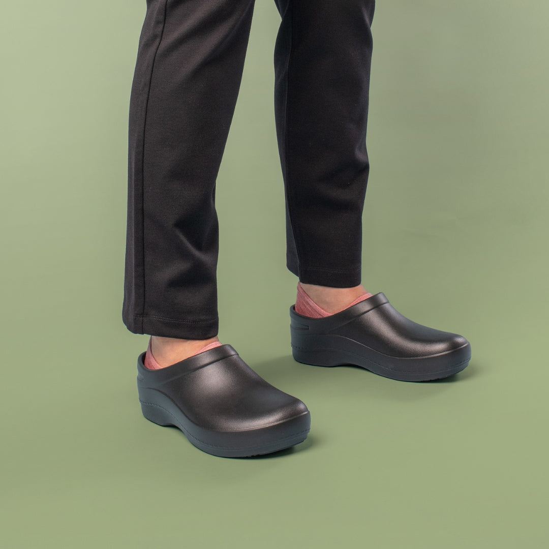 Black wipe-clean mules on a model wearing black pants. Green backdrop.