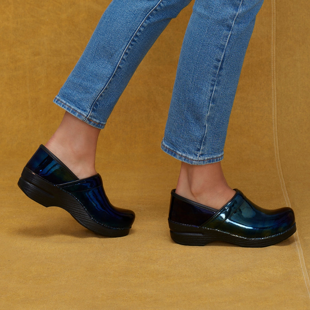 Black patent clogs on model wearing blue jeans. Mustard colored backdrop.