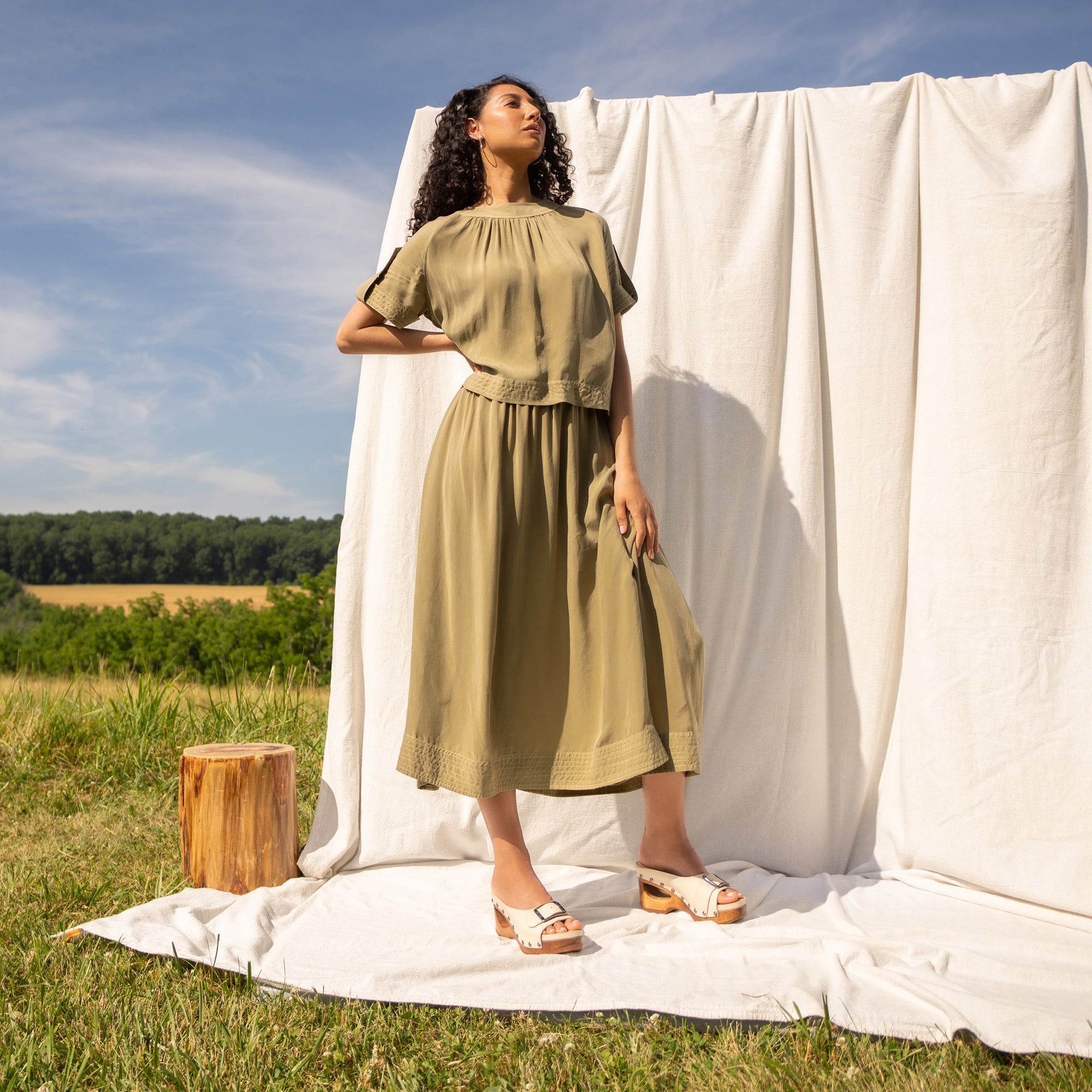 Woman in field standing in front of a neutral fabric backdrop wearing a khaki green outfit and cream wooden mules.