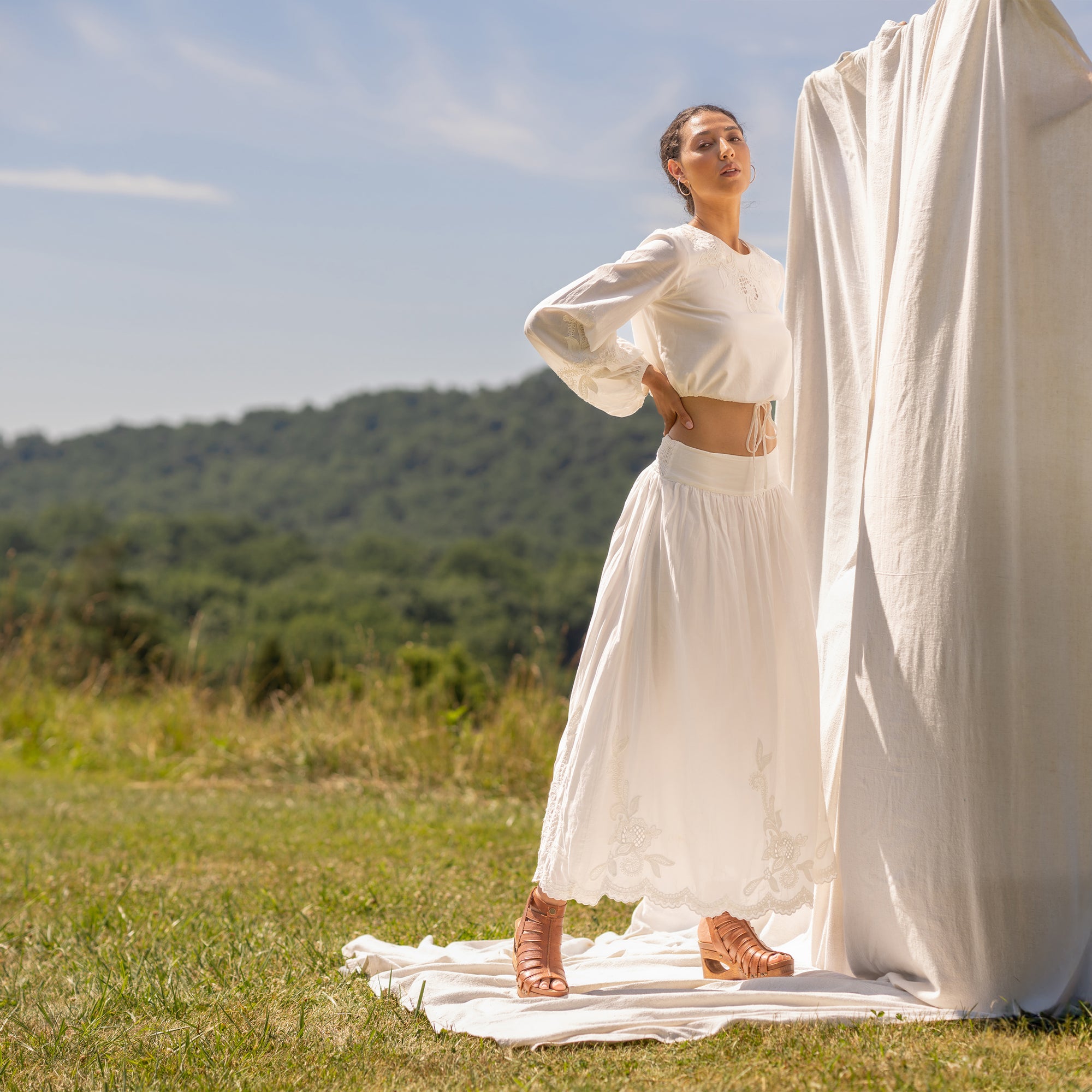 Woman standing in field in front of neutral backdrop in neutral outfit and tan strappy wood bottom sandals.