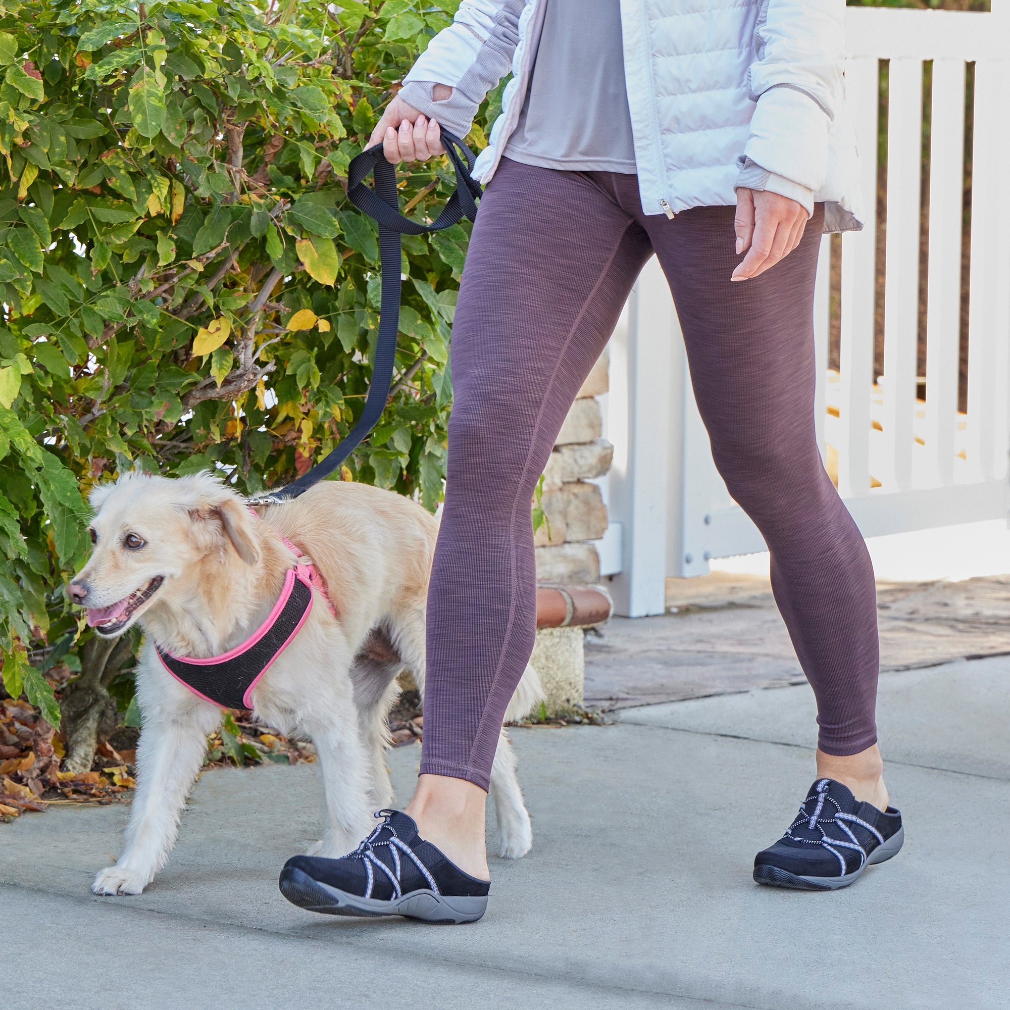 A woman wearing backless black sneakers and leggings while walking a dog.