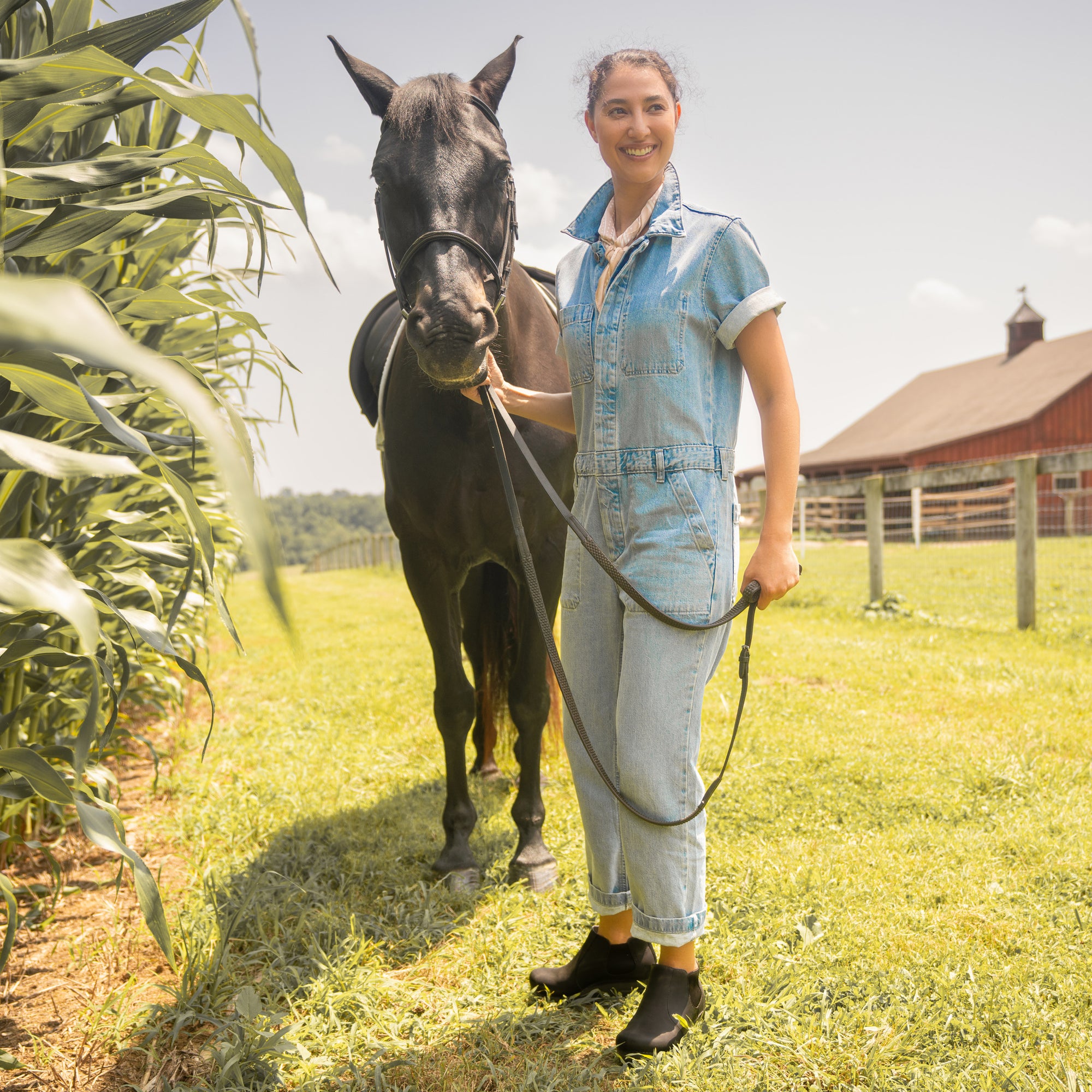 A farmgirl in denim coveralls wearing black boots while working with horses.