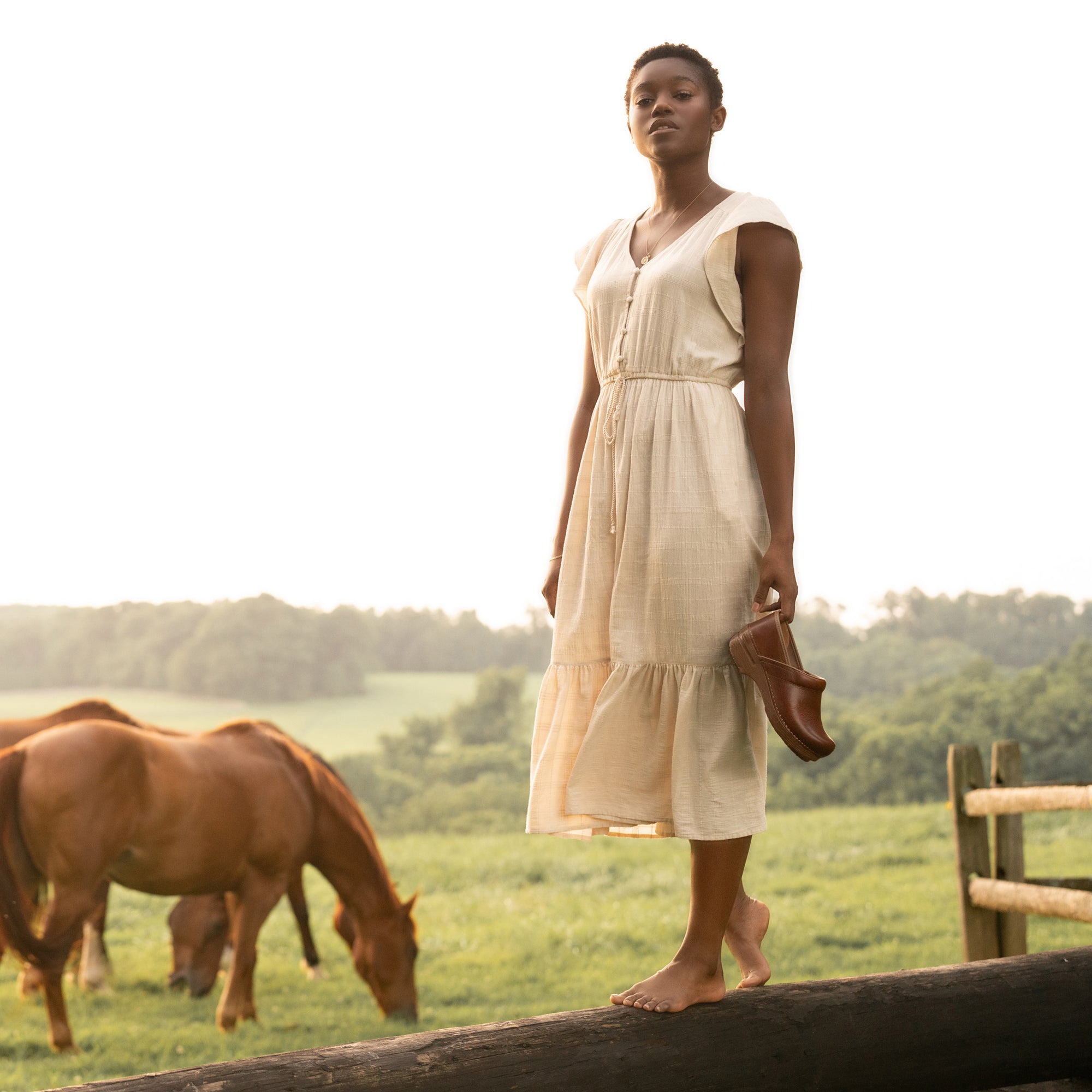 Woman holding dark brown clogs while walking on a log.