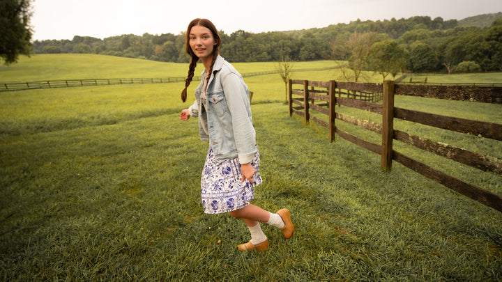A woman in a dress and denim jacket wearing tan clogs with white wool socks in an open field.