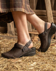 Image of a woman from the knee down in a plaid skirt and brown mules in a barn with straw.