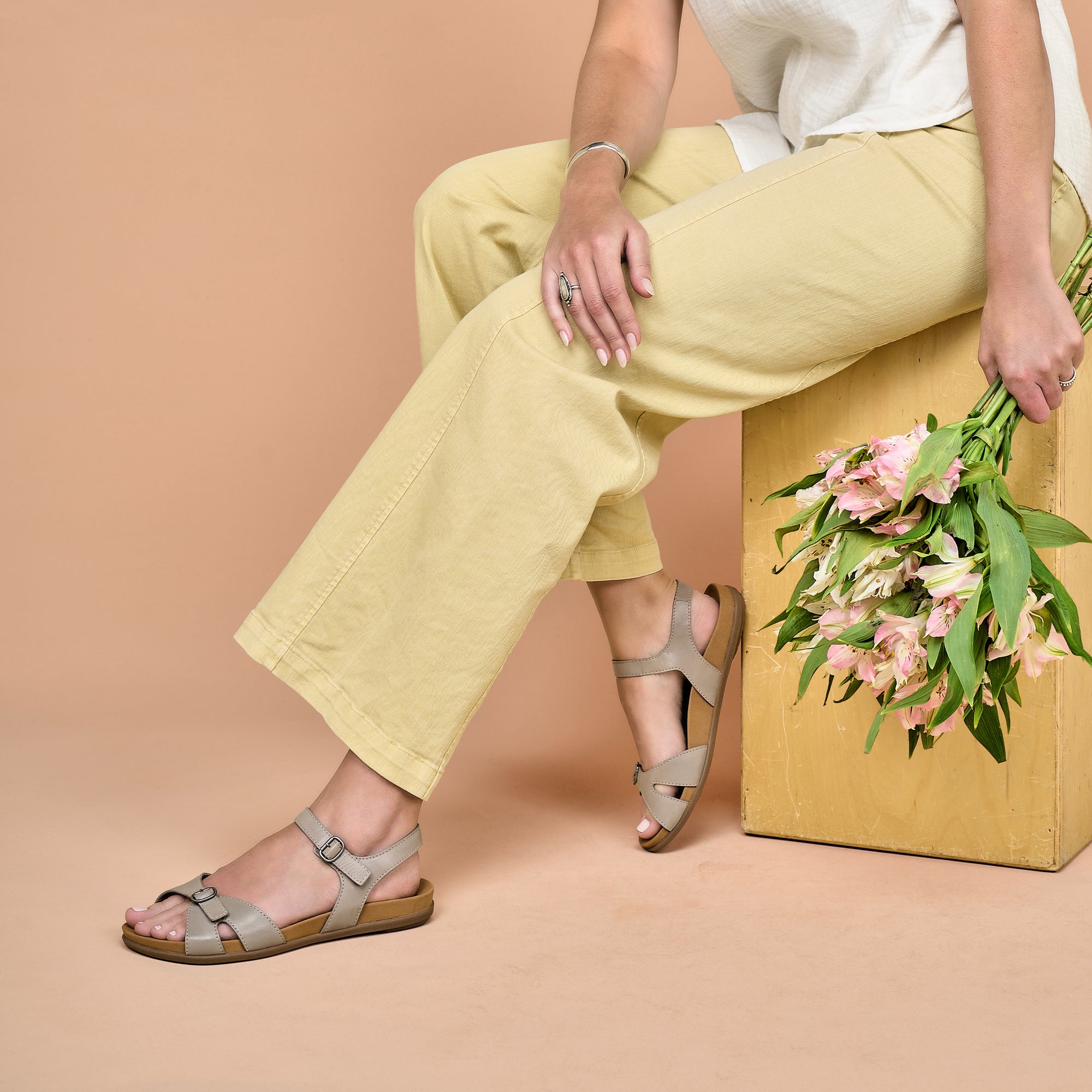 A woman sitting and holding flowers while wearing stone leather flat sandals.