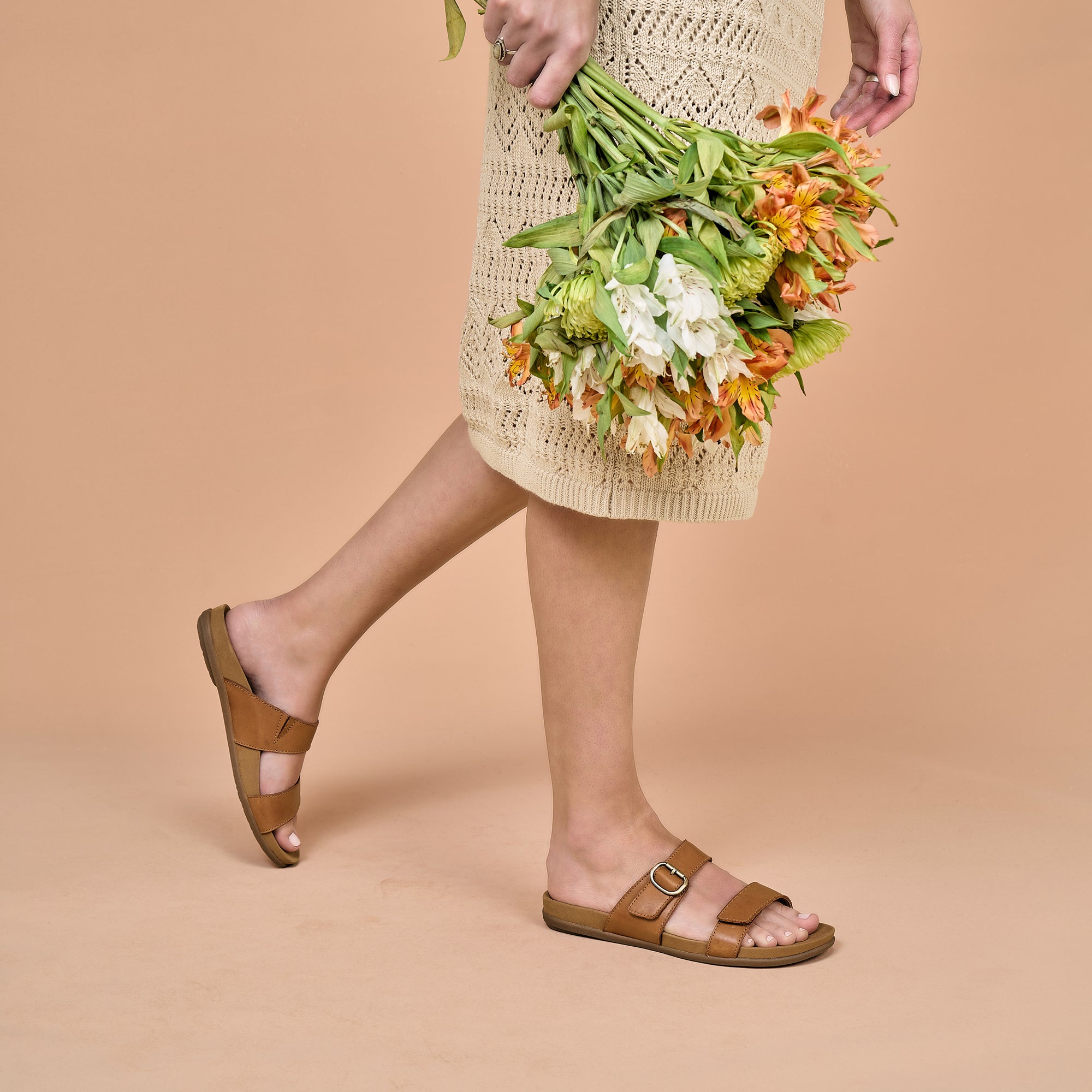 A woman in a dress holding flowers and wearing brown leather slide sandals.