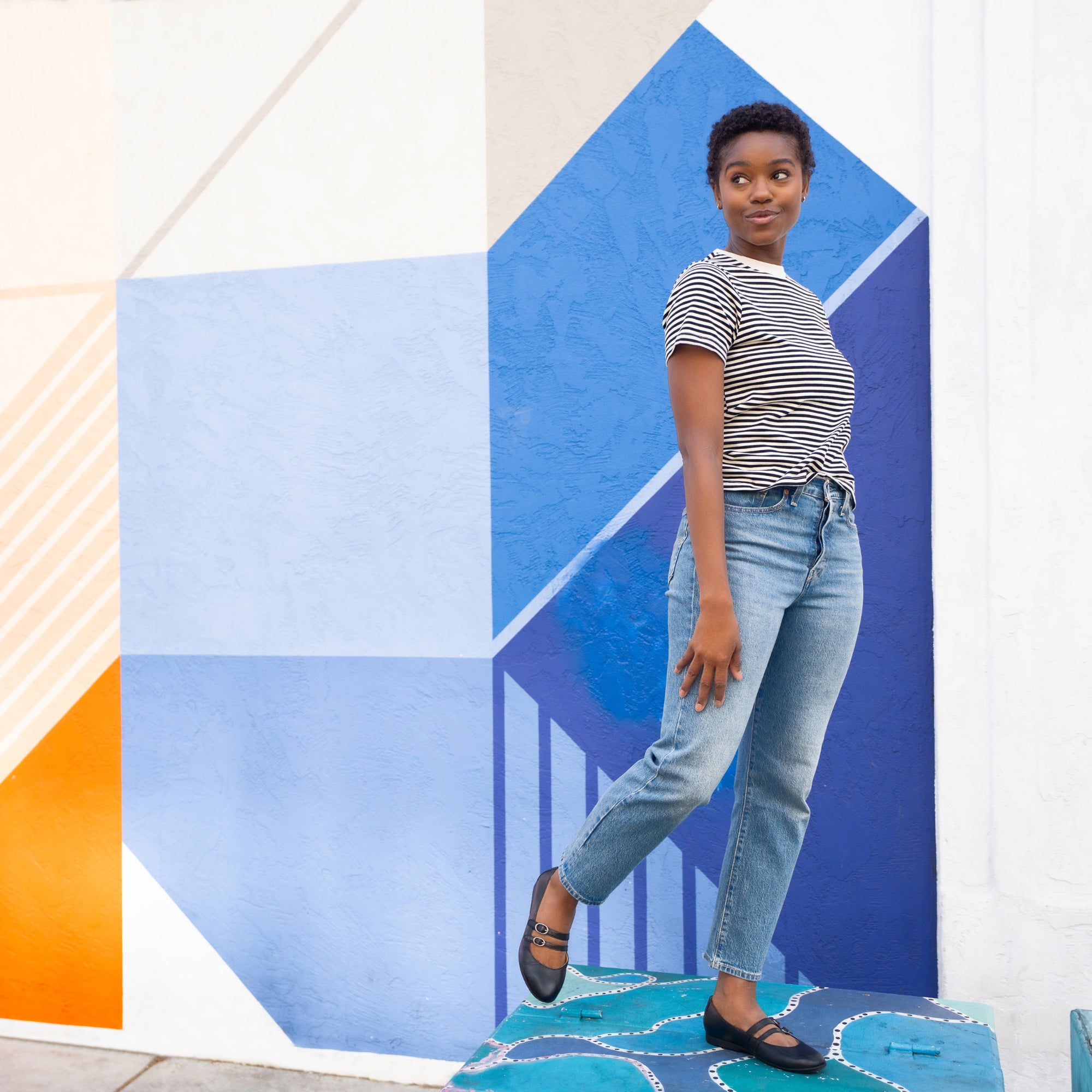A woman standing amongst street art wearing stylish, black two-strap Mary Jane flats.