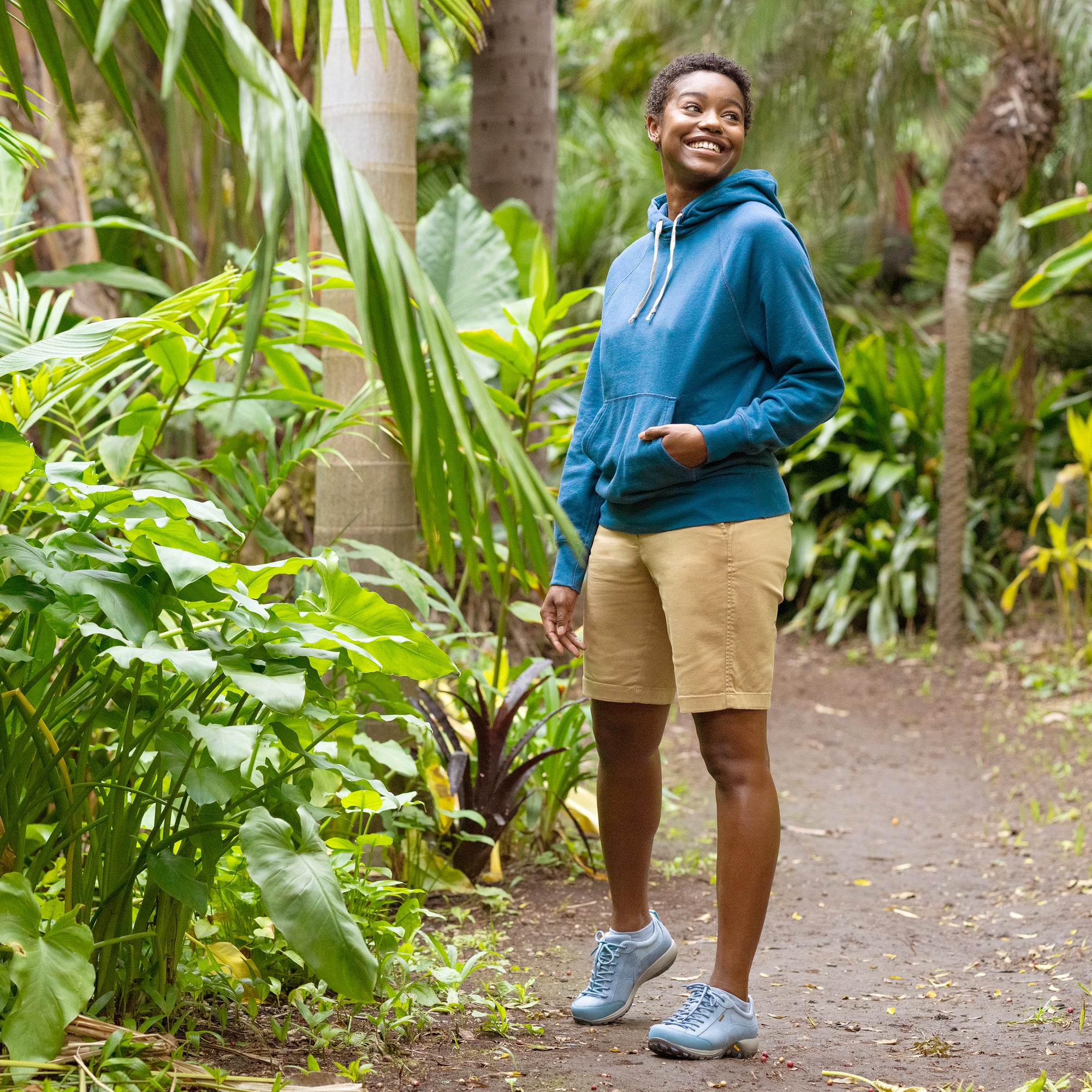 A woman standing with trees in a casual active outfit and sky blue outdoor sneakers.
