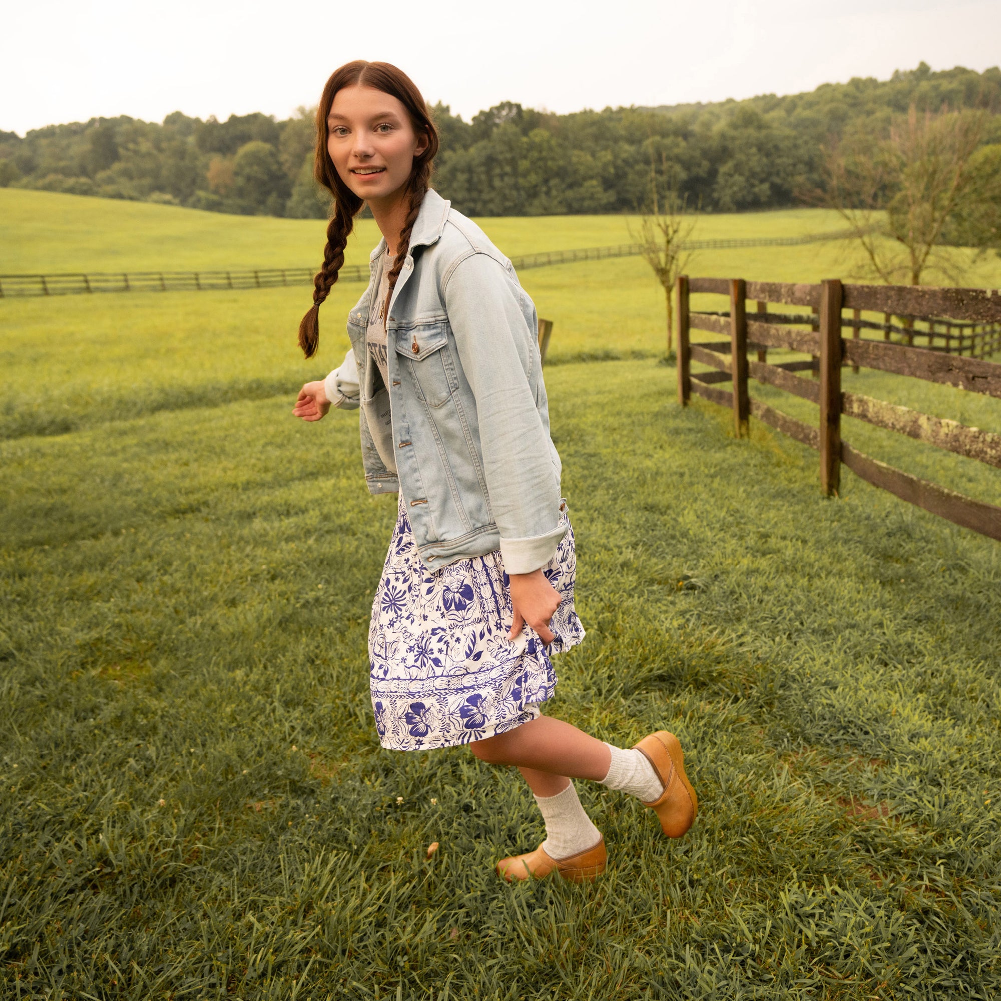 A woman with a dress and denim jacket wearing tan clogs with socks.