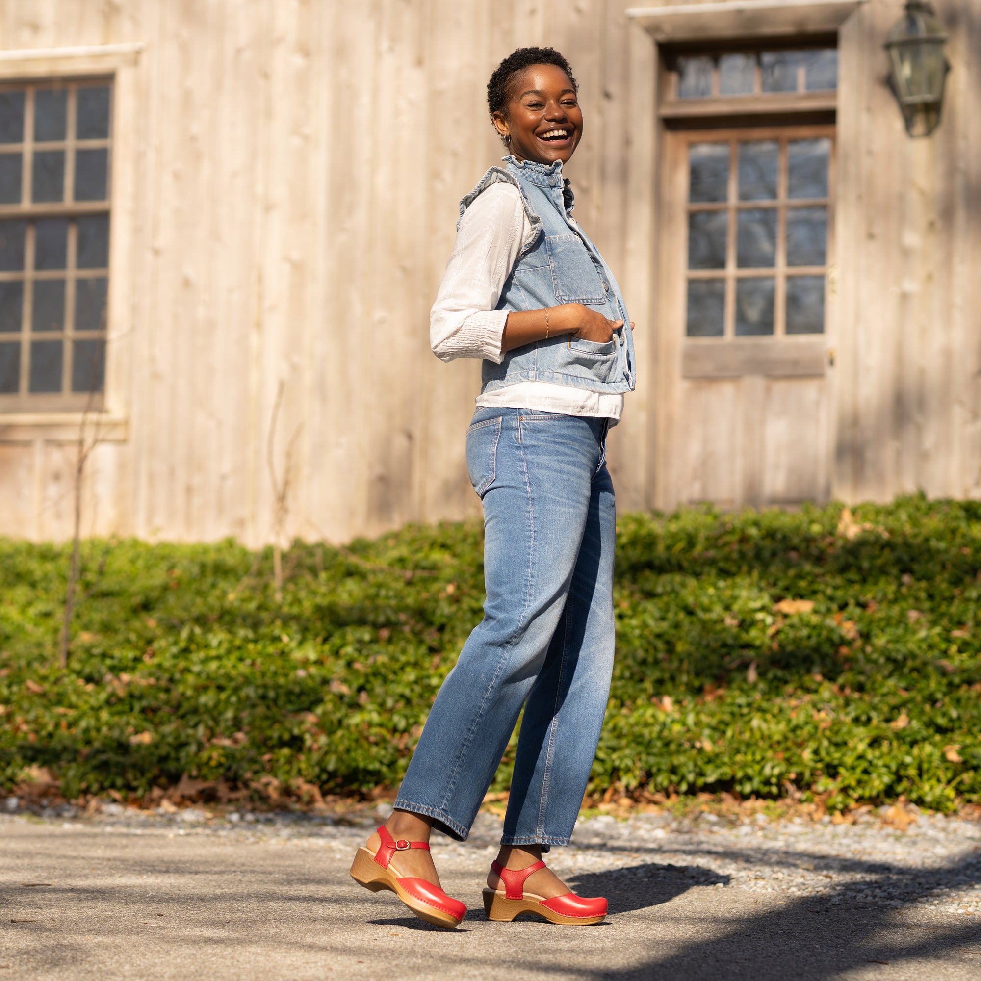 A woman smiling in the sun while wearing a denim outfit and red Mary Jane clogs.