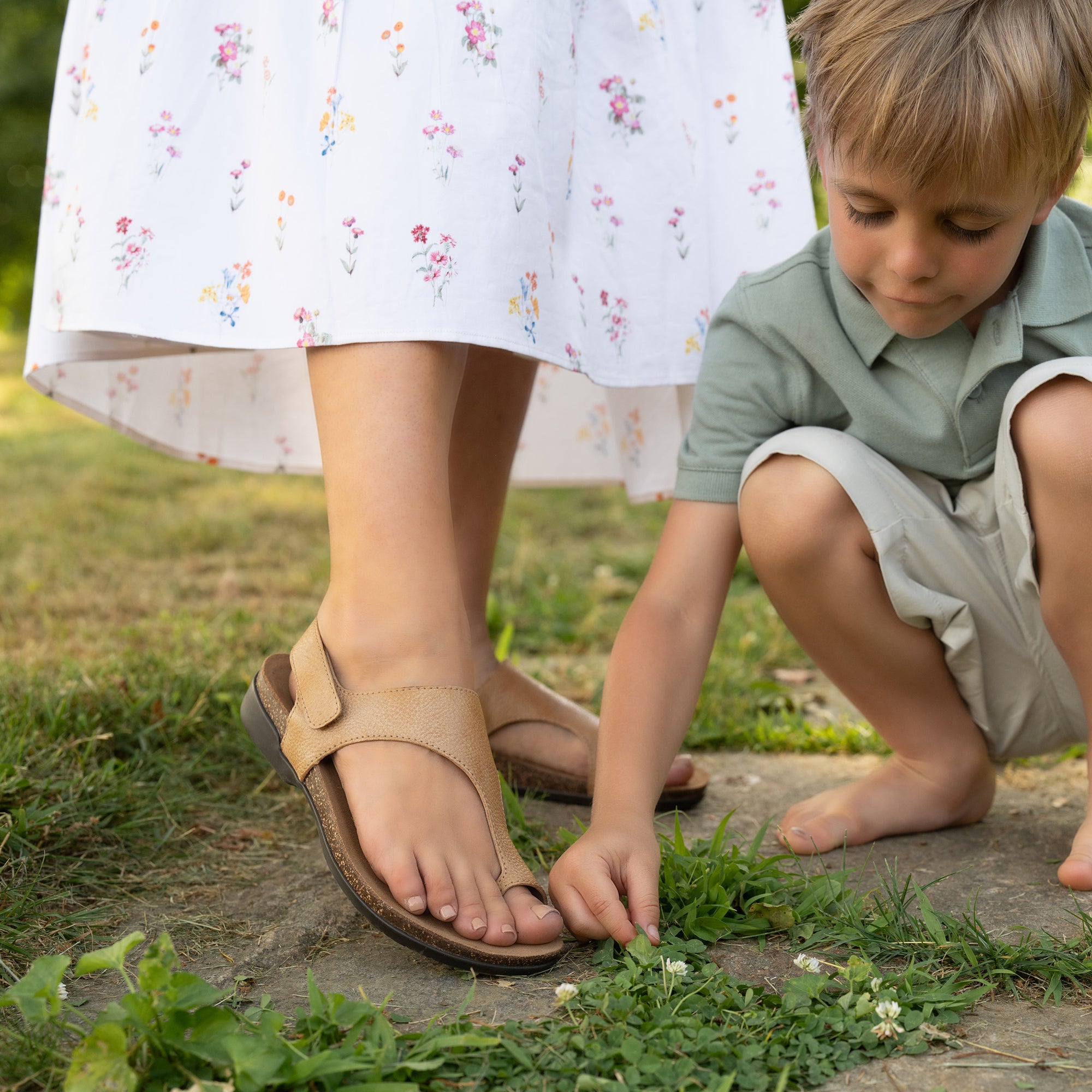Close up of a woman's tan sandals, showing only the bottom of her floral dress below the knee. Next to her, a young boy is crouched down playing with grass.