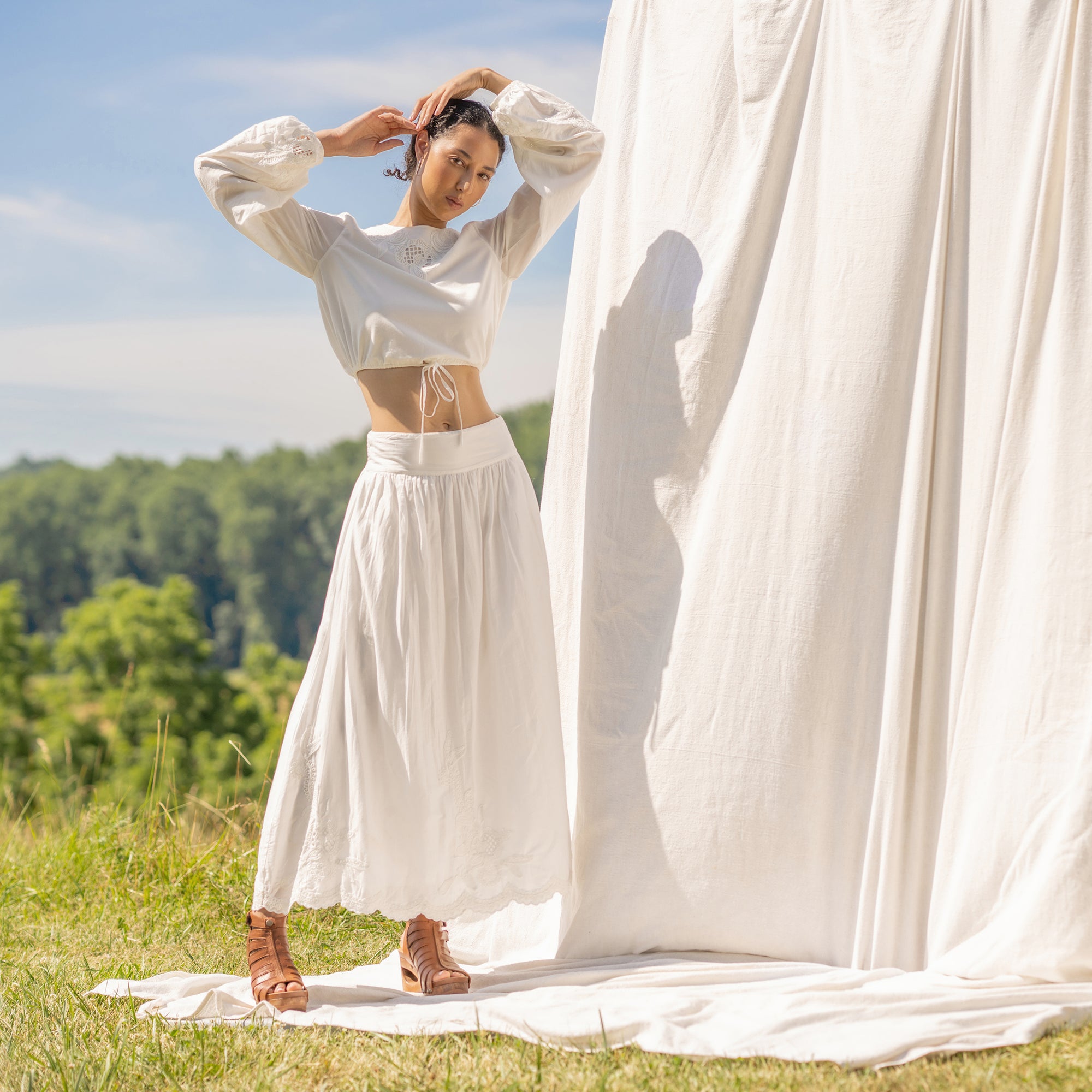 Woman standing in a field in front of billowy white linen. Woman is wearing a white cropped long sleeve shirt, long flowy white skirt, and tan wooden heels.