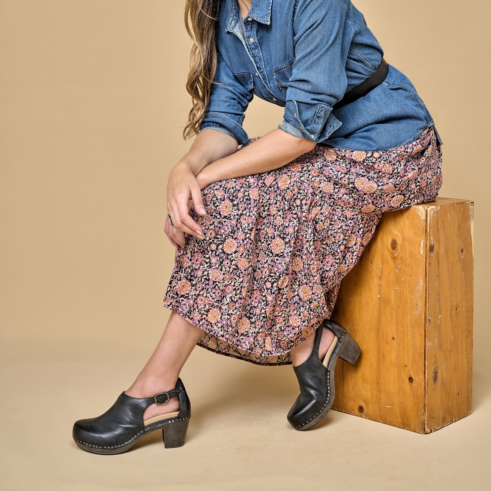 A woman sitting down on a wooden box wearing black leather heels with a stylish construction.