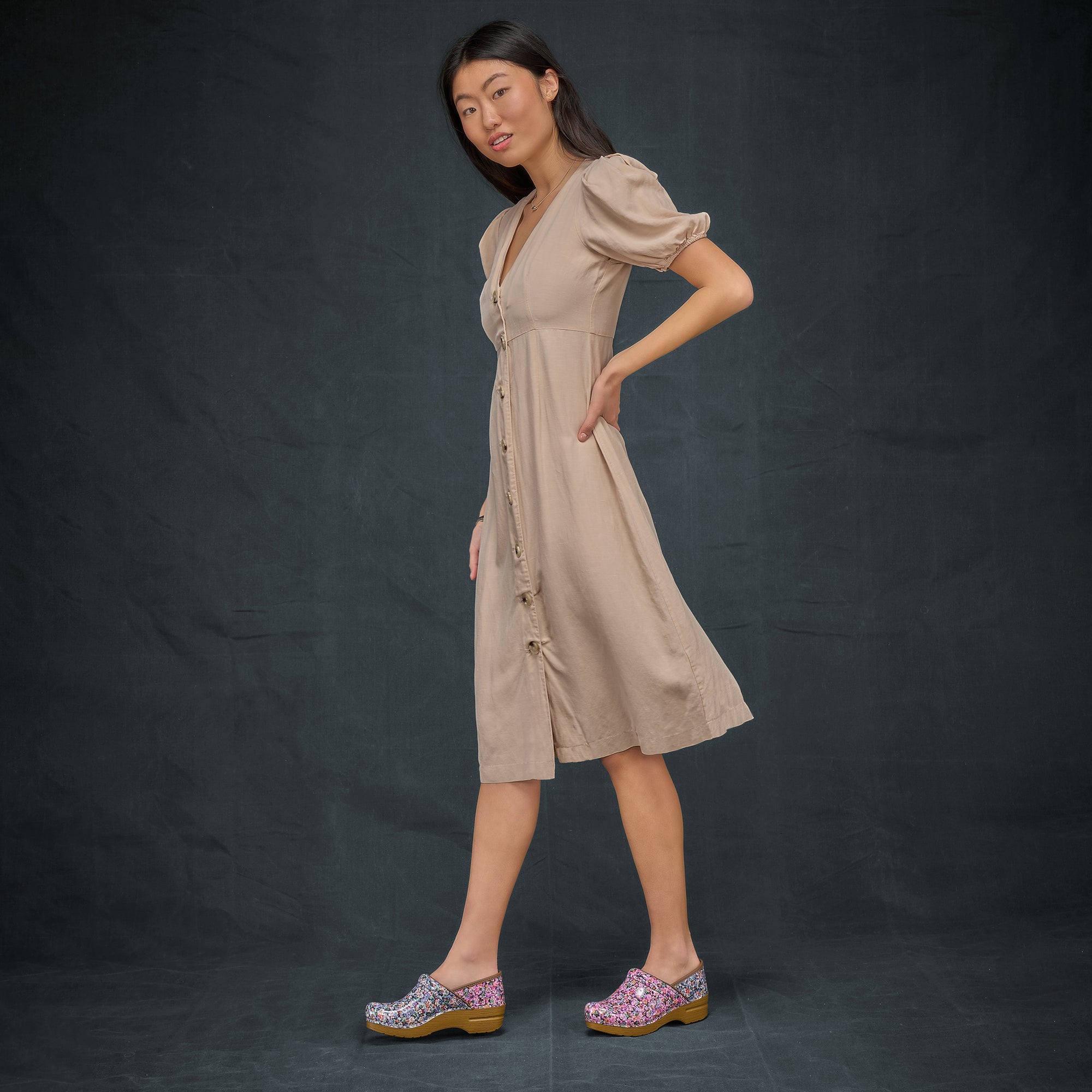 A woman wearing a tan dress and floral mismatched clogs in front of a black background.