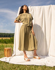 Woman in field standing in front of a neutral fabric backdrop wearing a khaki green outfit and cream wooden mules.