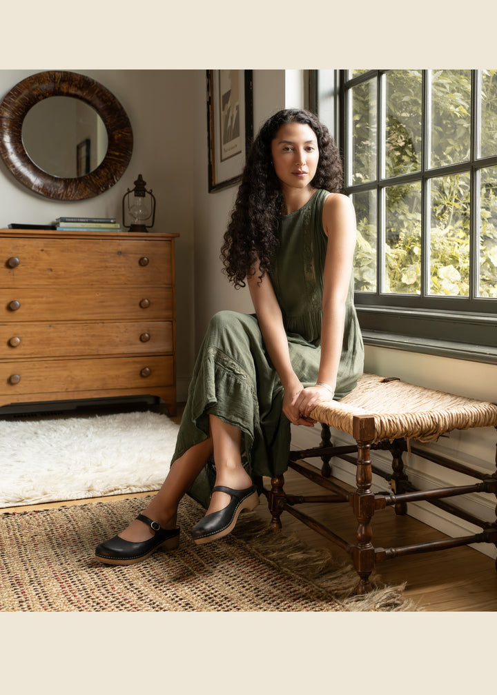 Woman with dark curly hair, green dress and black leather mary janes sitting on an indoor bench in front of a window.