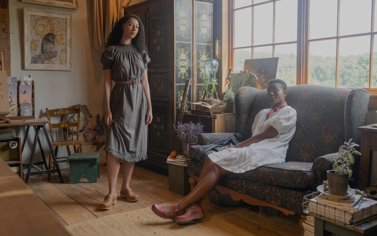 A woman in a gray dress stands in a rustic room while another woman in a white dress sits on an armchair by large windows.