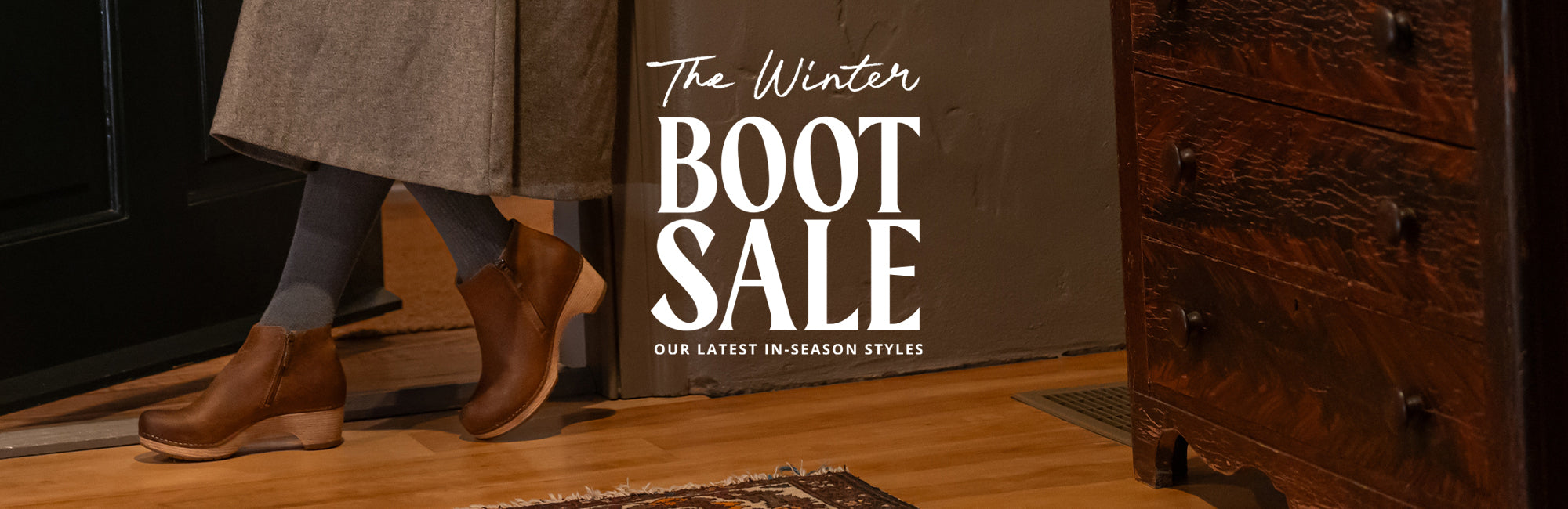 The left background shows waist down of a woman leaning up against a wall in a long skirt, tights, and brown boots. Towards the right of the background shows a dark wooden dresser. Center foreground text says "The Winter BOOT SALE. Our latest in-season styles."