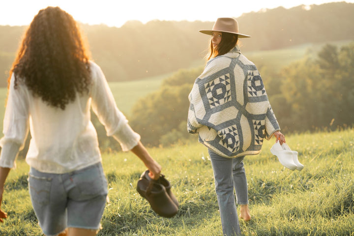 Girl carrying white clogs and another carrying brown backless clogs while walking through a meadow.