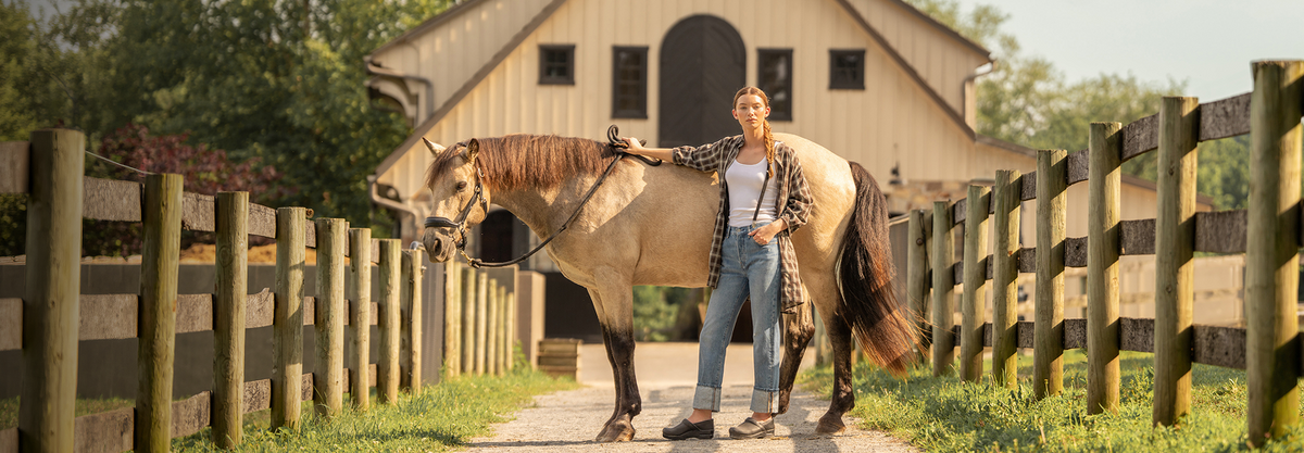 A woman stands on a path between wooden fences next to a light brown horse, with a barn in the background. She is wearing a plaid shirt, jeans, and Dansko clogs.
