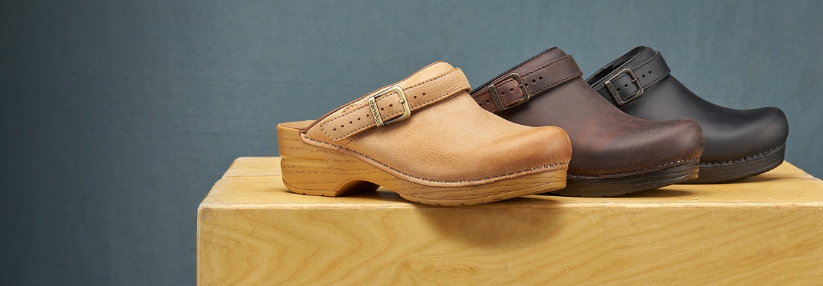 Three Dansko clogs in tan, dark brown, and black, arranged on a light wood block against a dark blue-gray background.