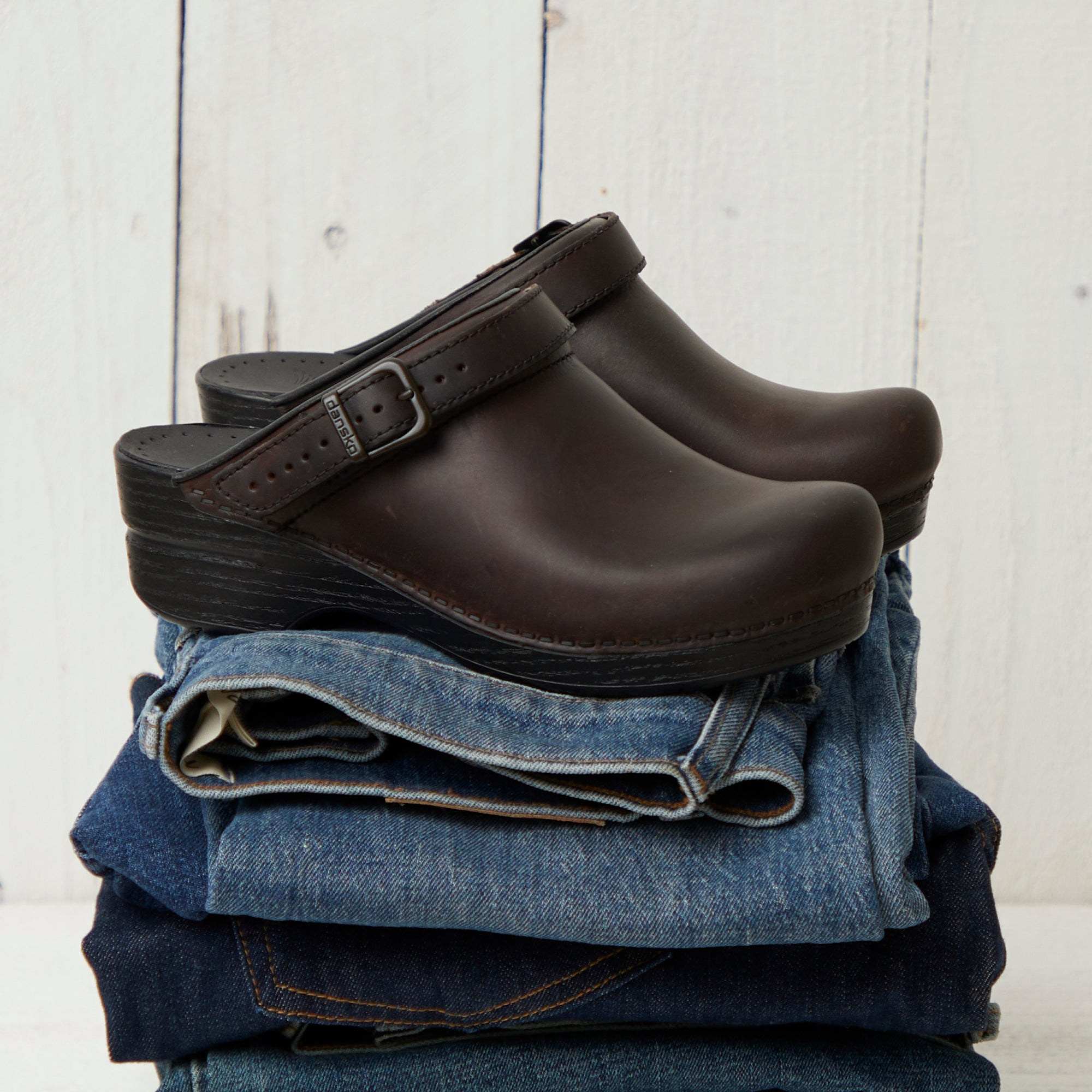 Brown backless clogs with an adjustable strap stacked on top of pairs of jeans.