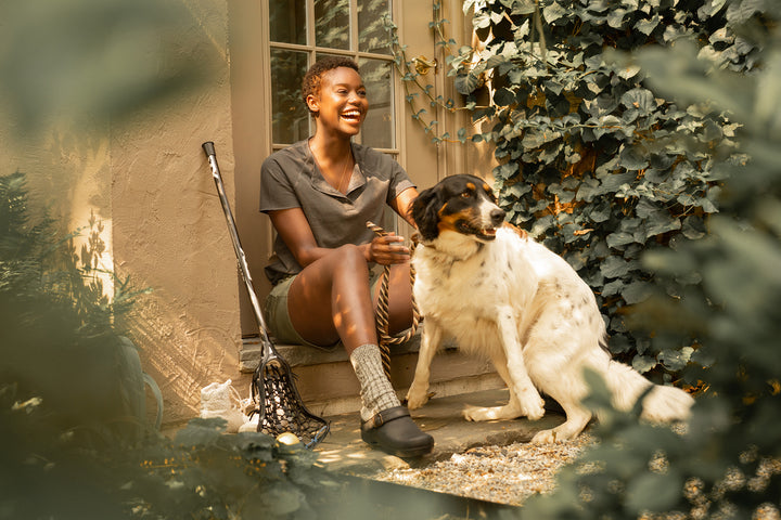 Woman with a dog wearing brown backless clogs and smiling widely.
