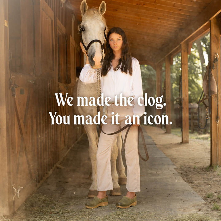 Lifestyle image of a brunette girl in white long sleeve shirt, khaki pants and Professional Green Burnished clogs standing in front of a white horse with the exterior of a barn in the background. Text reads: We made the clog. you made it an icon.