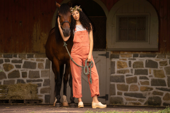 A woman in orange overalls and gorgeous tan clogs standing in a stable with a horse.