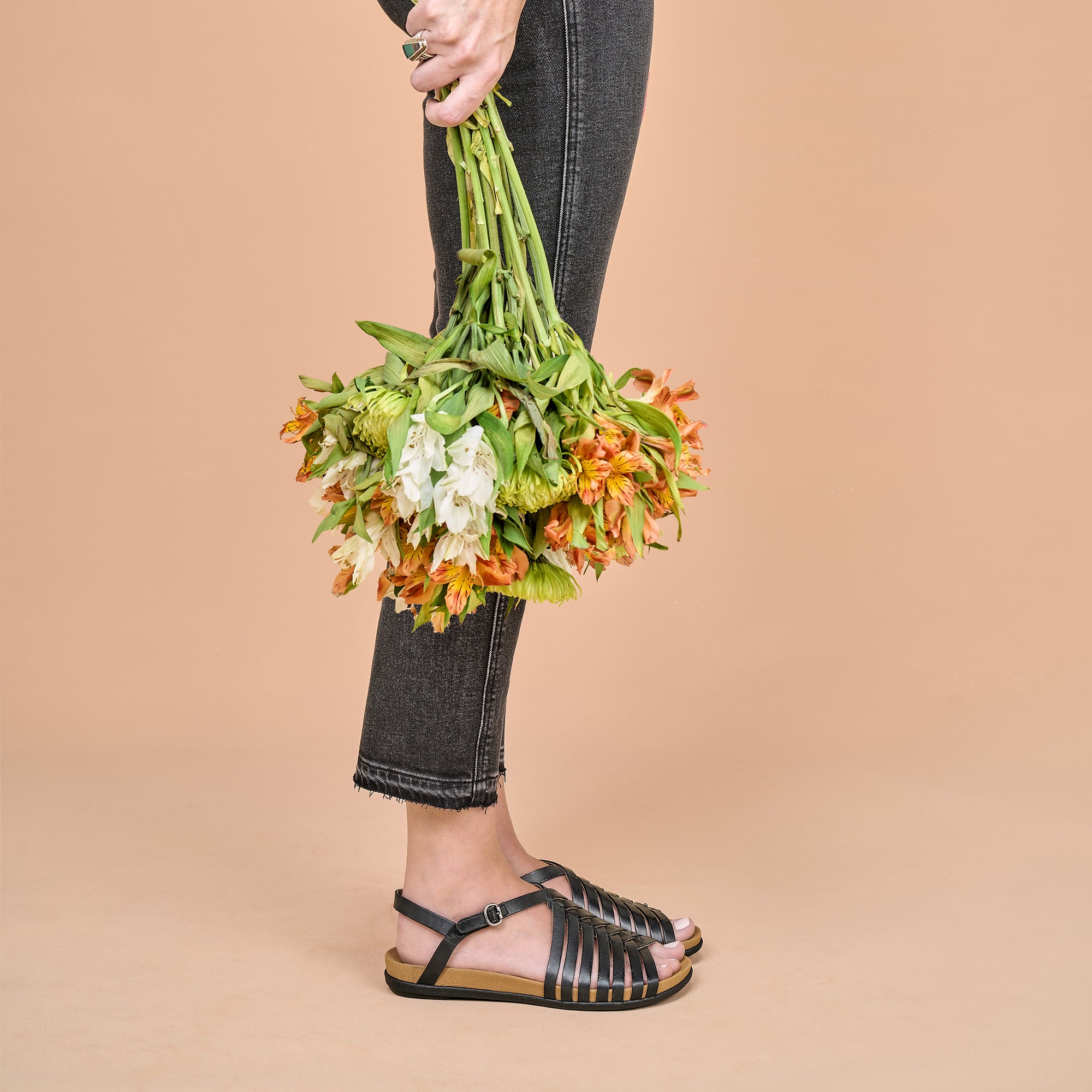 A woman holding flowers and wearing black fisherman sandals against a pale orange background.