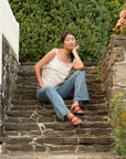 Woman sitting on outdoor stone steps wearing a neutral tank top, light blue jeans, and red flat strappy sandals.