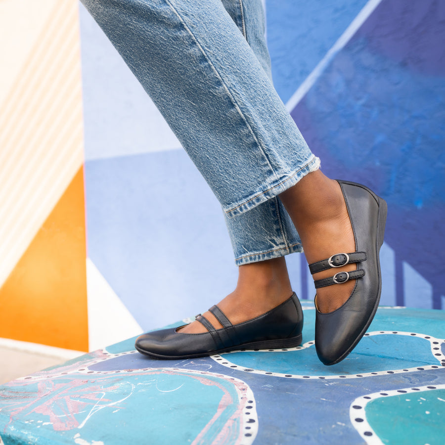 A clos up of a woman's stylish two-strap Mary Jane flats while standing amongst street art. 