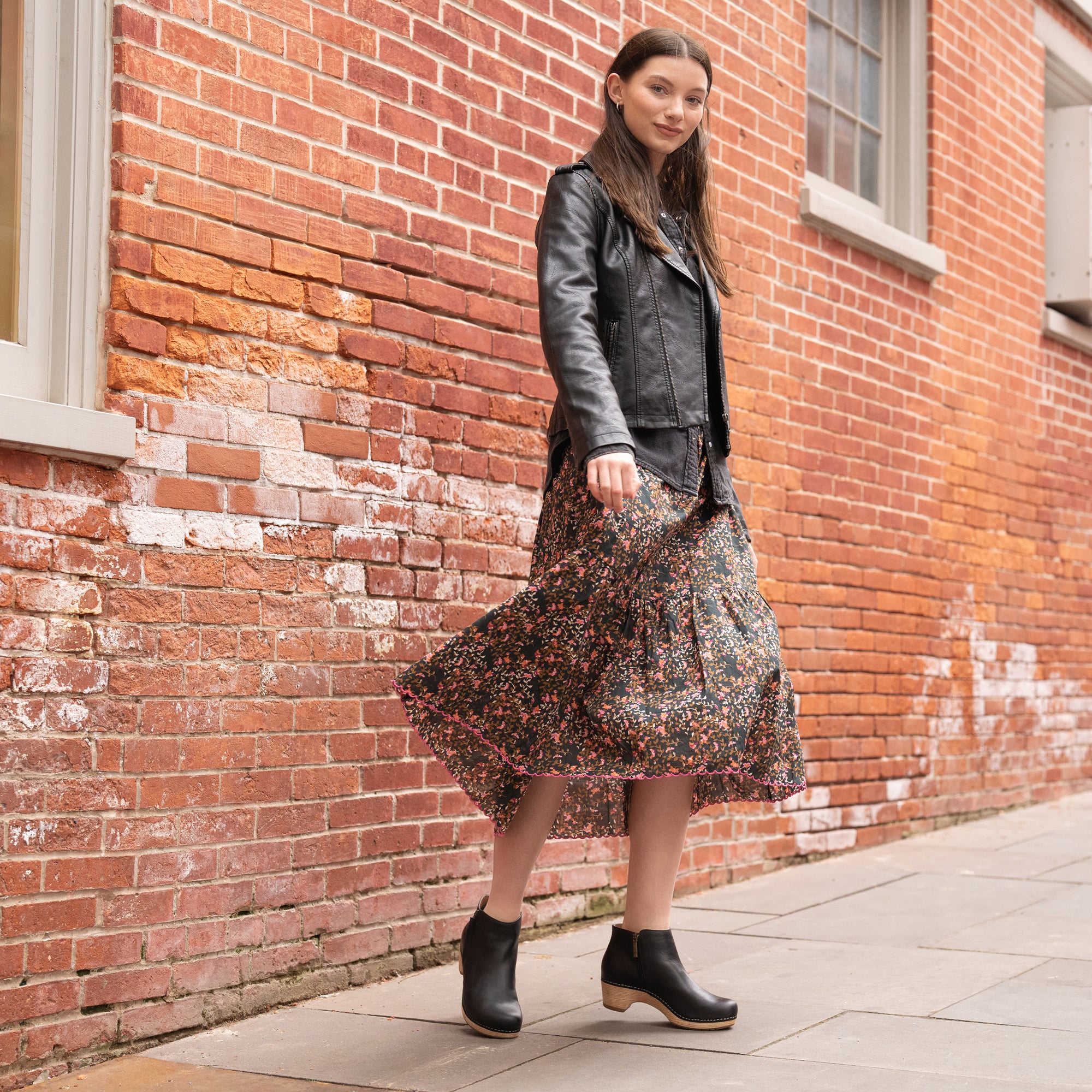 A woman in a stylish leather jacket and floral dress wearing black leather booties with a faux wood sole.