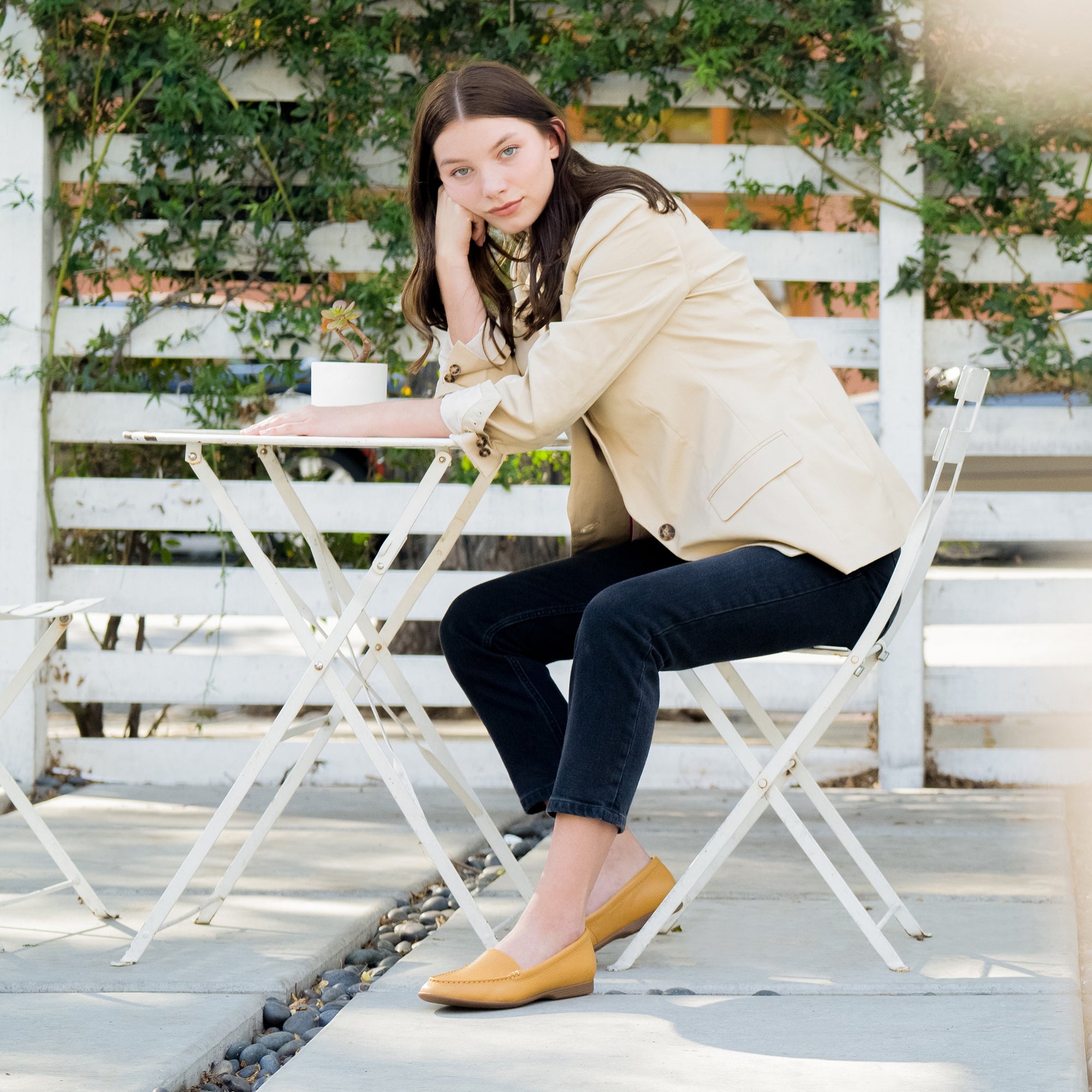 A woman sitting in a garden cafe wearing mustard loafer flats and work attire.