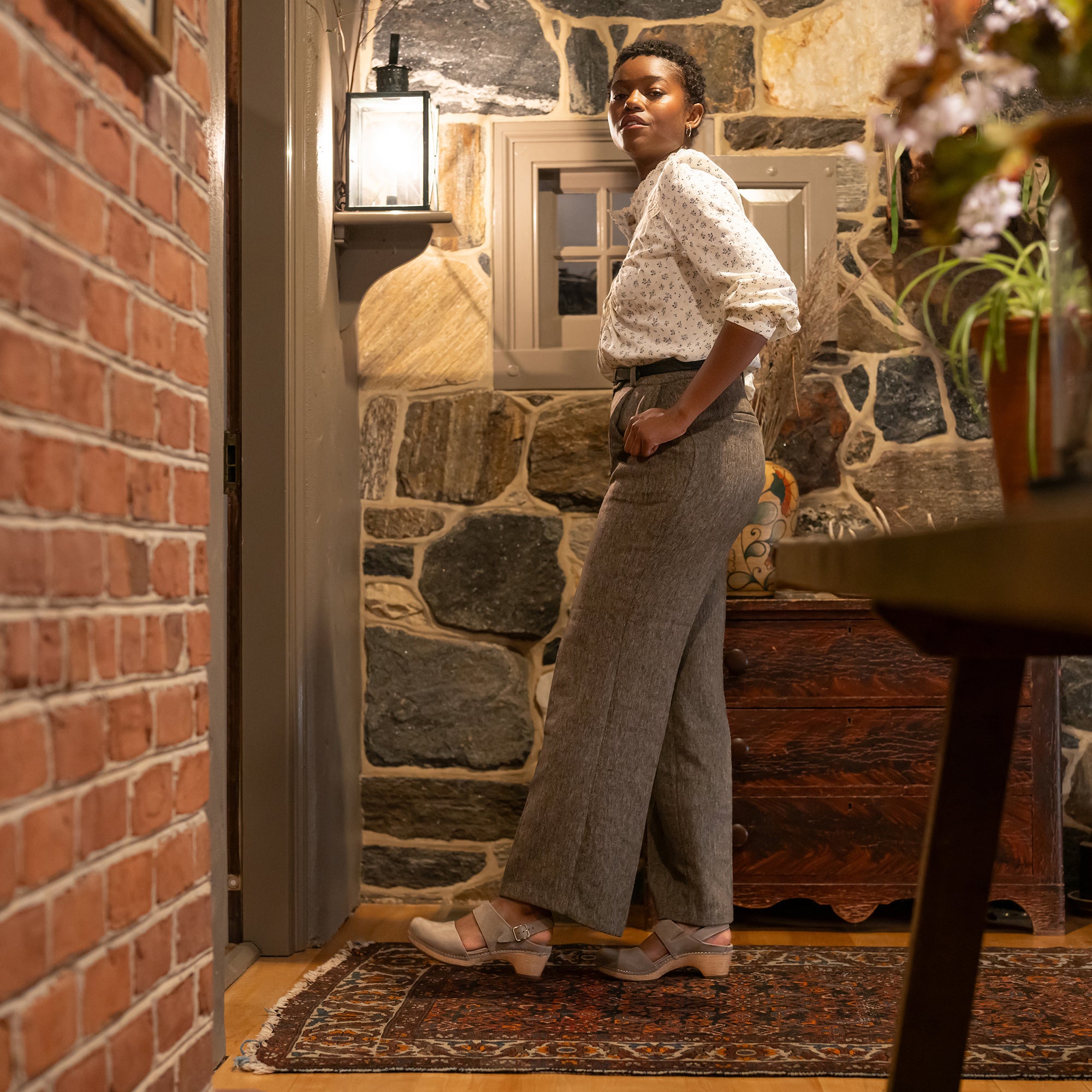 A woman in a stylish outfit wearing light grey transitional sandals against a rustic backdrop.