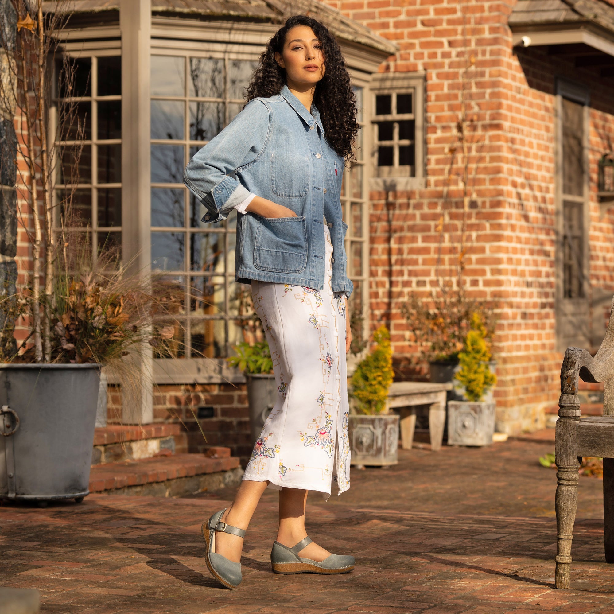 Woman standing in front of brick building wearing a denim jacket, white patterned dress and blue mary janes.