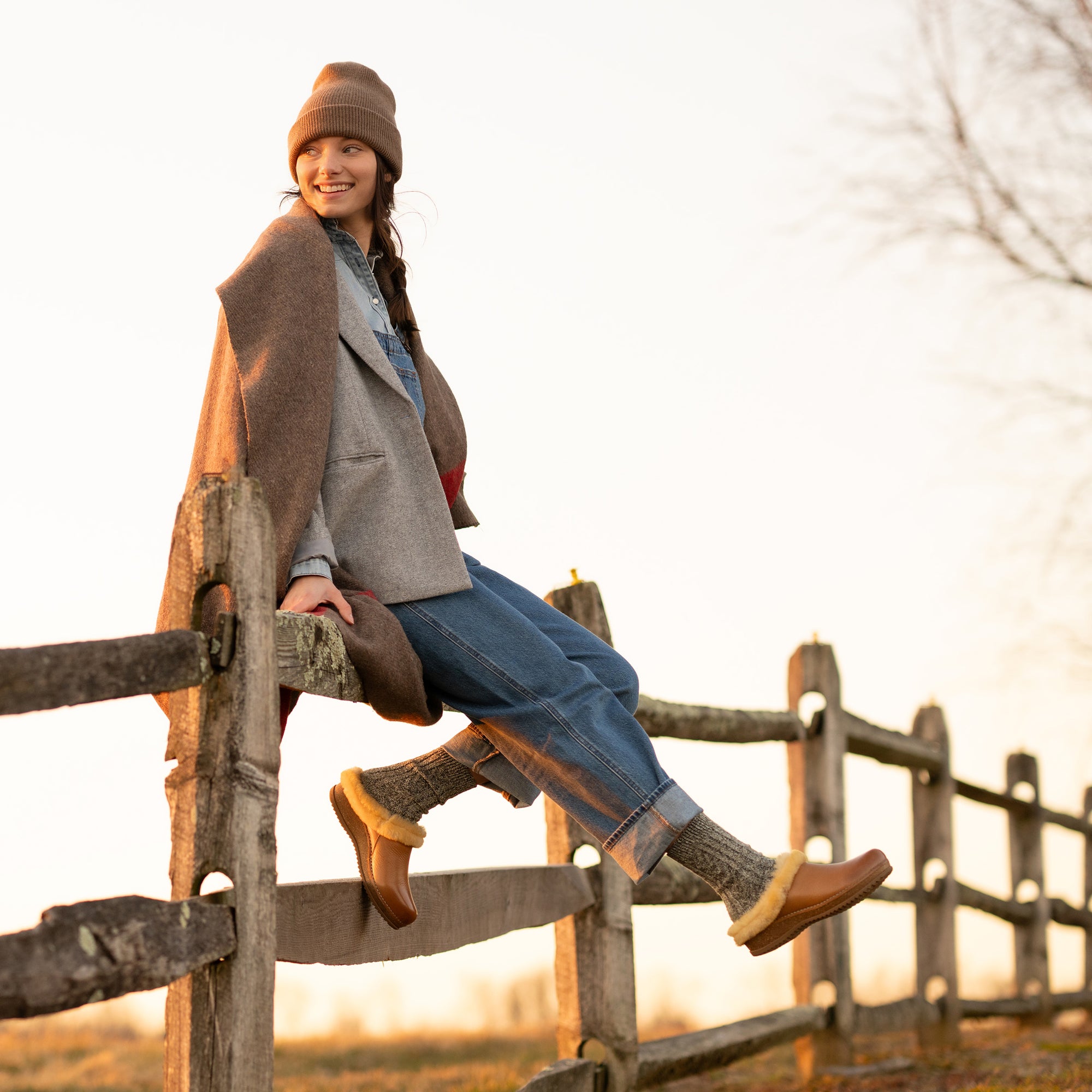 A woman sitting on a fence while wearing shearling-lined mule clogs.