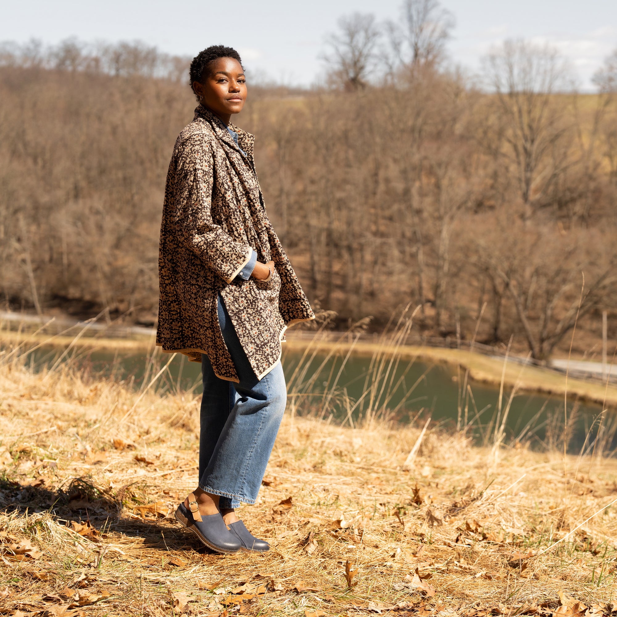 A woman wearing a quilted coat and jeans with blue leather clogs that stand out in a fall field.