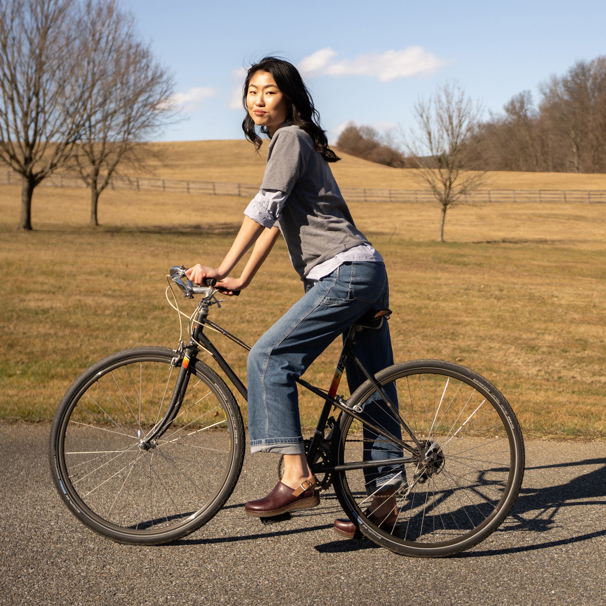 A woman in autumn attire and cordovan clogs riding a bike.