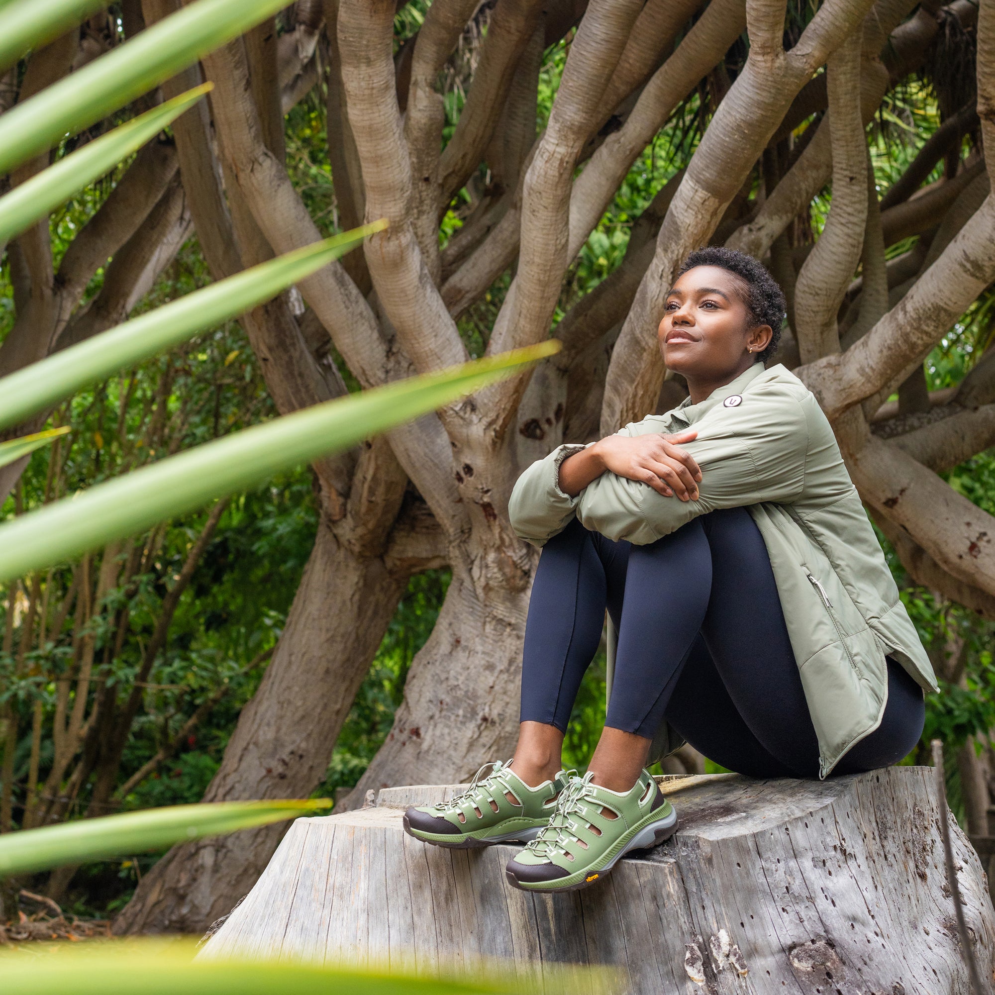A woman sitting on a tree stump in nature wearing lightweight and durable green sneakers.