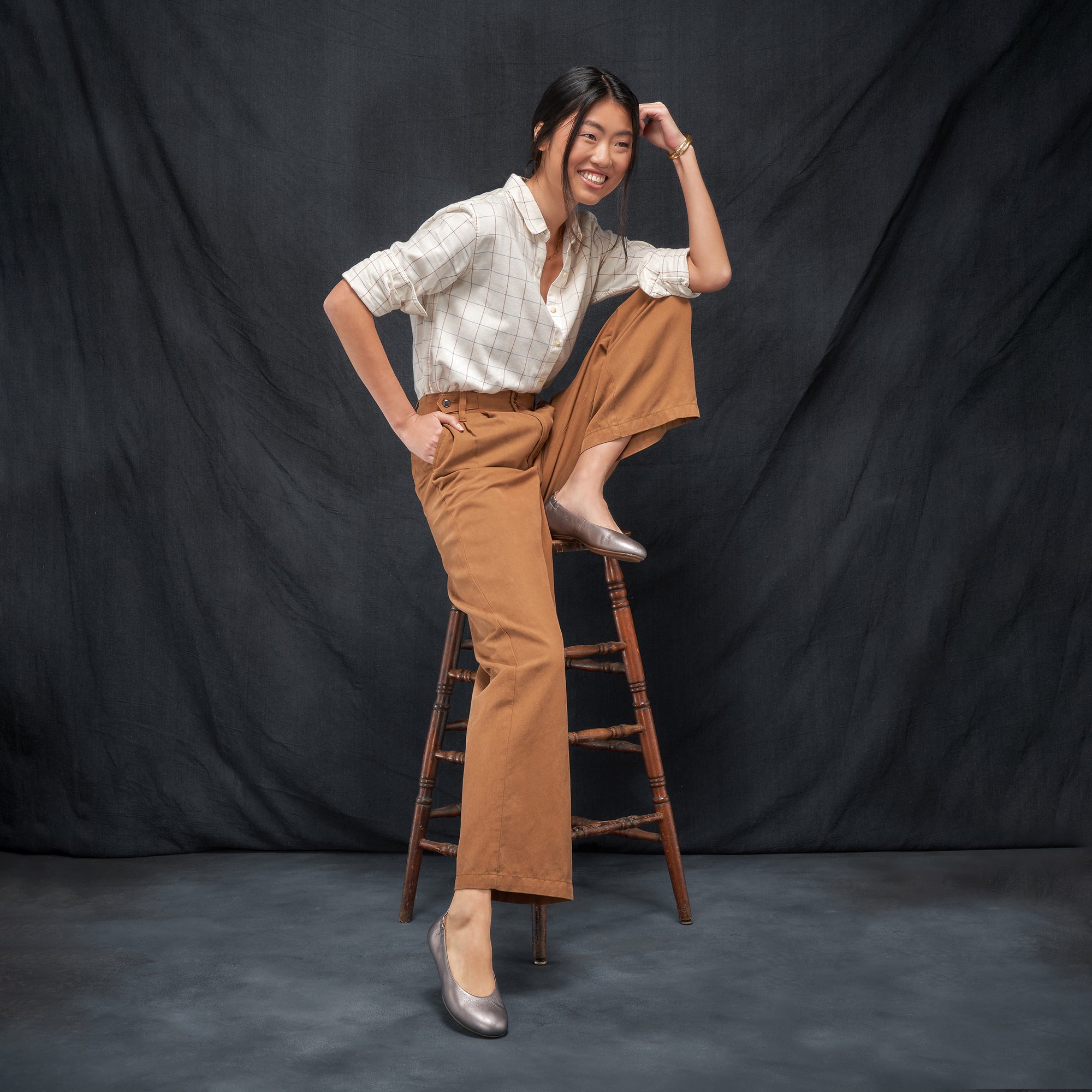 A woman sitting on a stool in a stylish outfit wearing metallic silver ballerina flats.