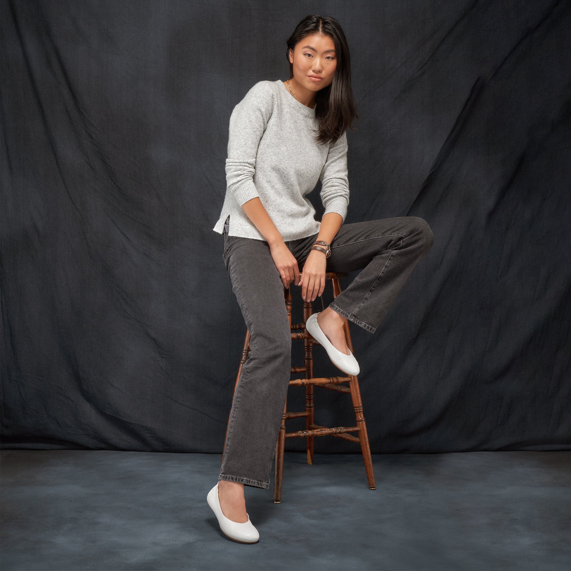 A woman sitting on a stool in a stylish outfit wearing white ballerina flats.