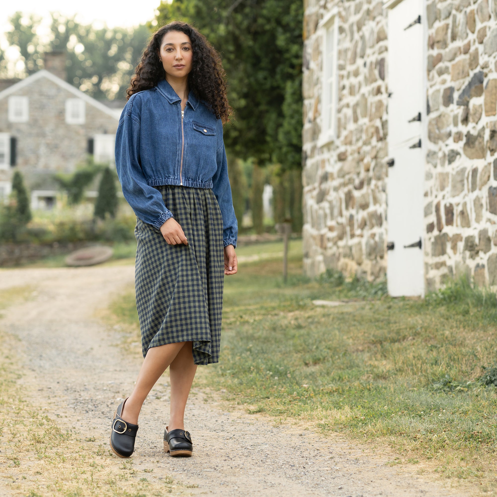 A woman in a plaid dress and denim jacket wearing black clogs with a buckle.