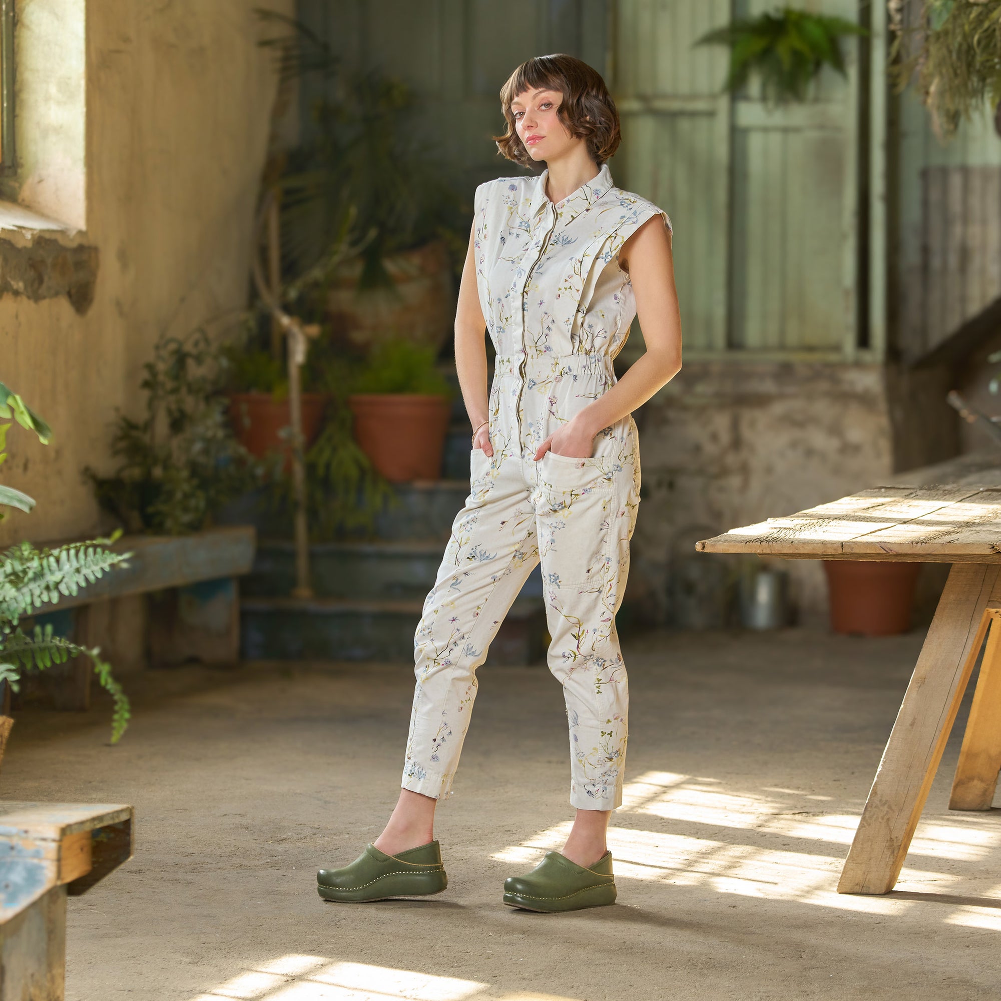 Woman in industrial space with greenery wearing a floral jumpsuit and green platform Dansko clogs.