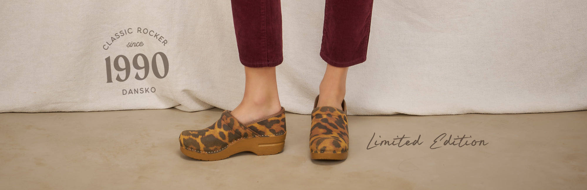 Beige background showcasing below the knee of a woman standing in the foreground. She is wearing dark pants and leopard print clogs. On the left side of the image, there is a logo that reads "Classic Rocker since 1990 Dansko" and to the right there is copy that reads "Limited Edition".