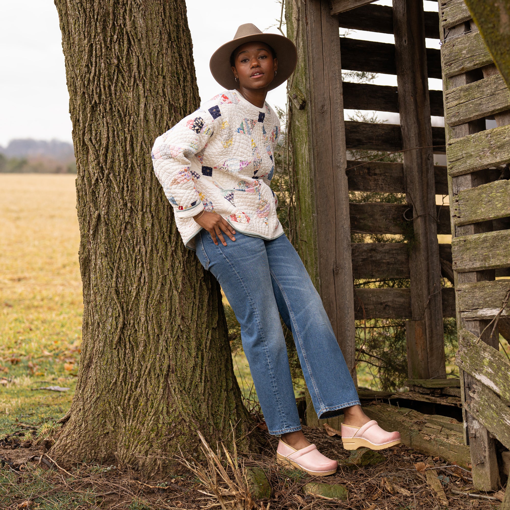 A woman on a farm in a quilted sweater wearing brightly neutral pink clogs.