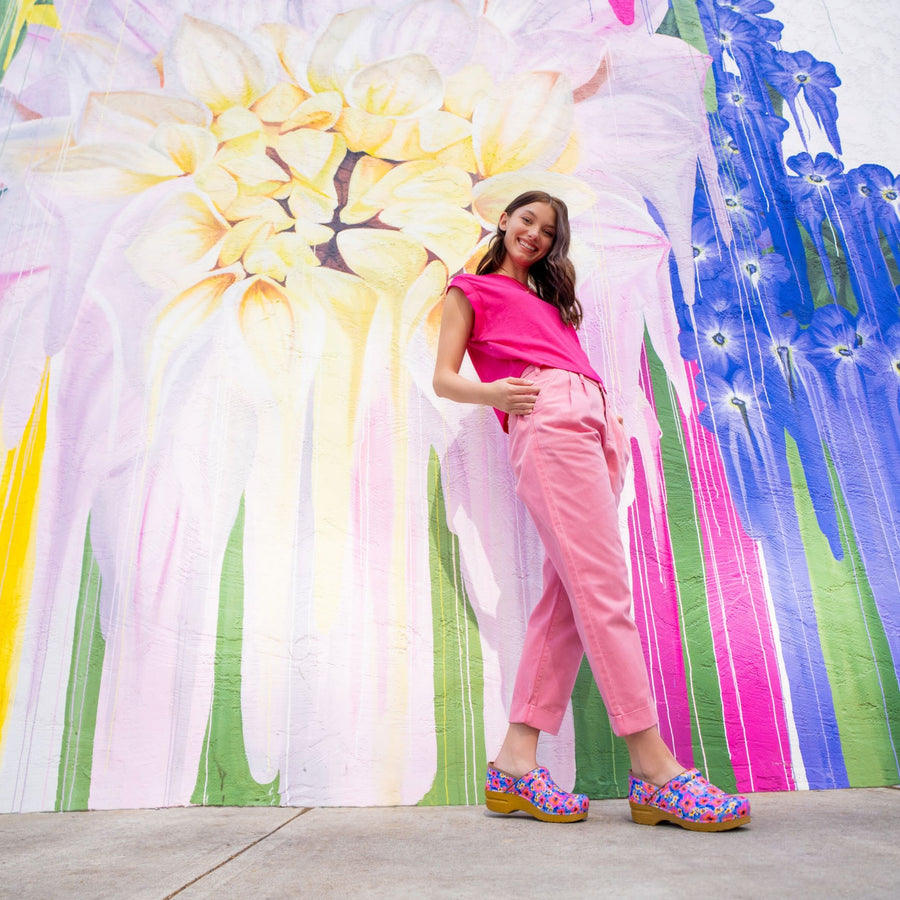 A woman wearing all pink standing in front of a mural wearing flowery patent clogs.