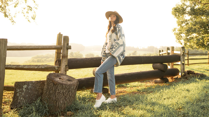 A woman in an eccentric quilted jacket and a wide brim hat standing in a field while wearing supportive white clogs.