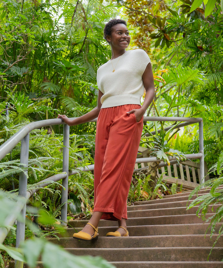 A woman in a white shirt and flowy pants standing with plants while wearing yellow Mary Jane sandals.